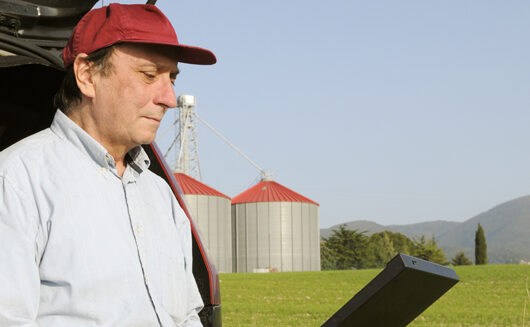 Man in red cap using laptop in a rural field, with silos and distant mountains in the background.