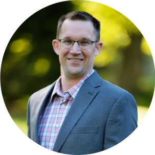 A man wearing glasses, a plaid shirt, and a gray suit jacket is smiling outdoors with greenery in the background.