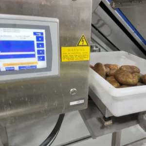 A plastic container filled with potatoes sits on a scale next to a control panel with an extendag and a warning label about electrical shock.