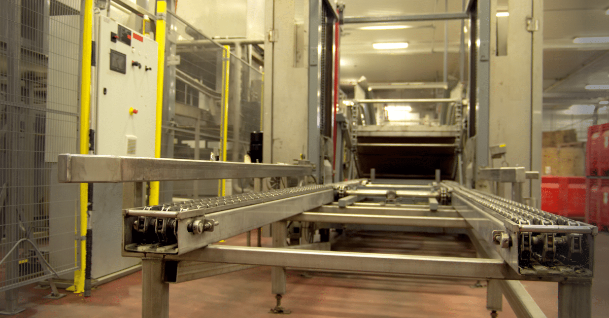 Conveyor system inside a factory with metal tracks, control panel on the left, and industrial equipment in the background.